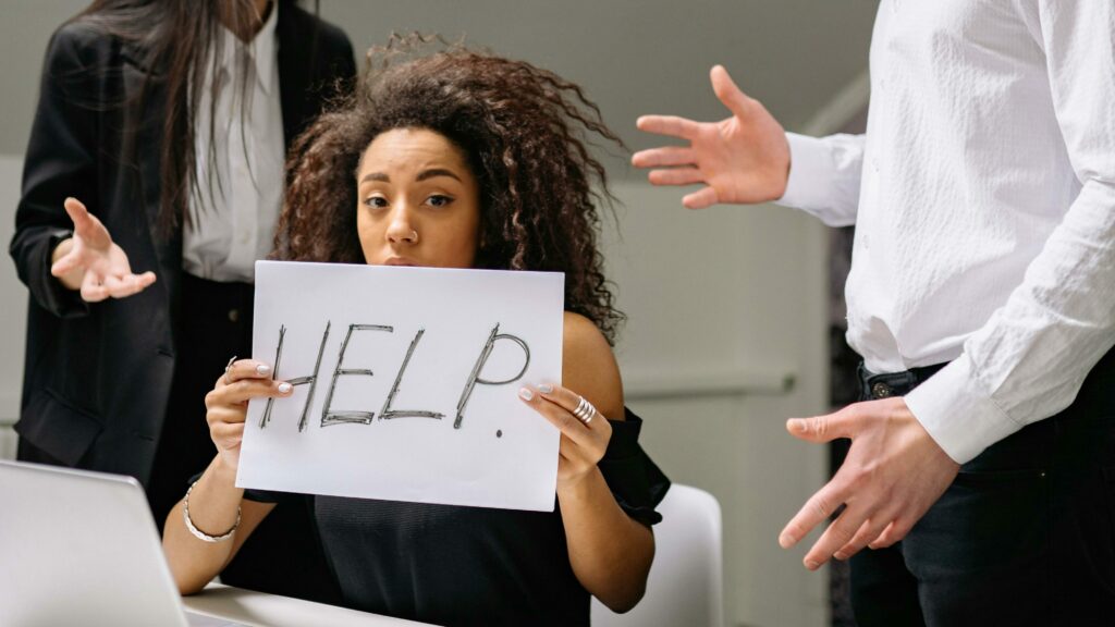 Woman holding HELP sign surrounded by colleagues in a tense office situation.